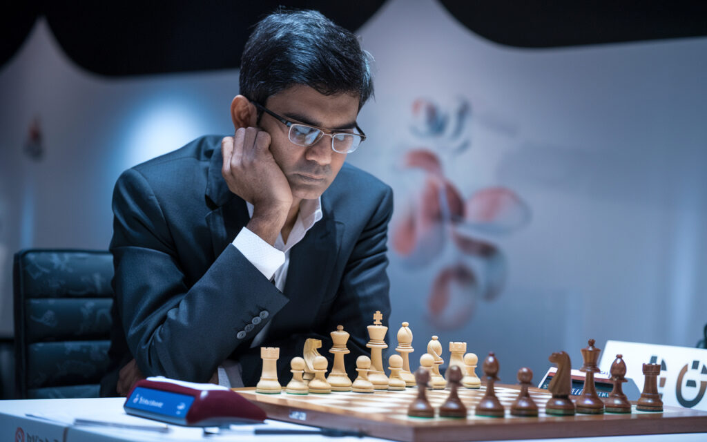 A photo of Viswanathan Anand, the maestro of Indian chess. He is sitting at a table with a chessboard. The board has a position from a game. The background is blurred and has a few chess pieces. The top left corner contains the headline "Viswanathan Anand: The Maestro of Indian Chess".