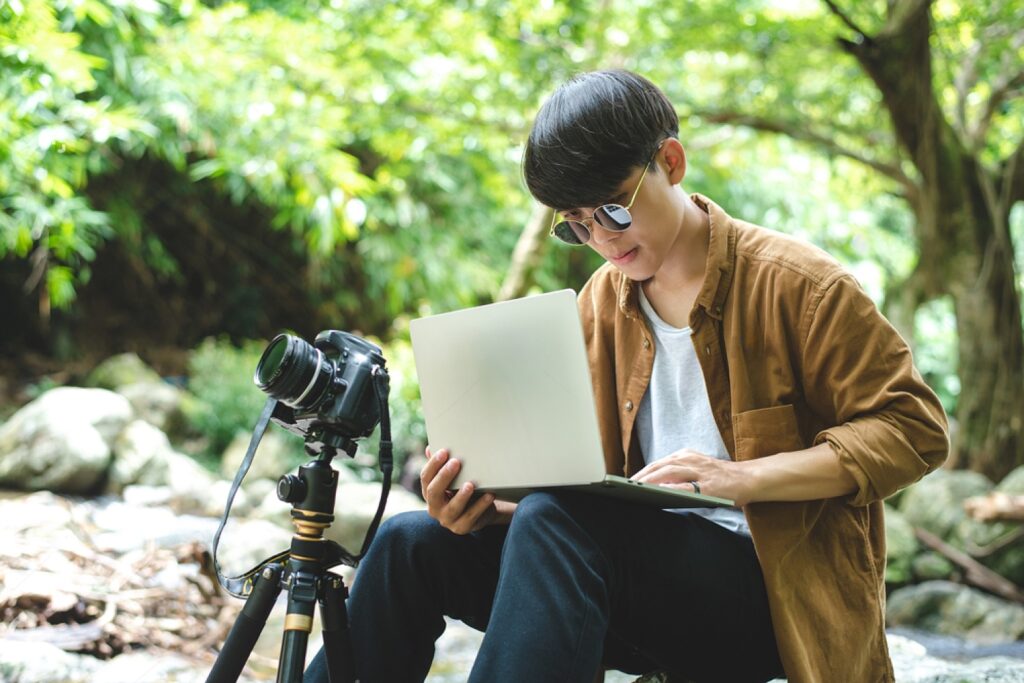 Asian man traveler photographer are taking photo and use laptop at beautiful forest. Travel photography, blogger, vlogger, camping, work aboard, adventure lifestyle, hipster, tourism concept.